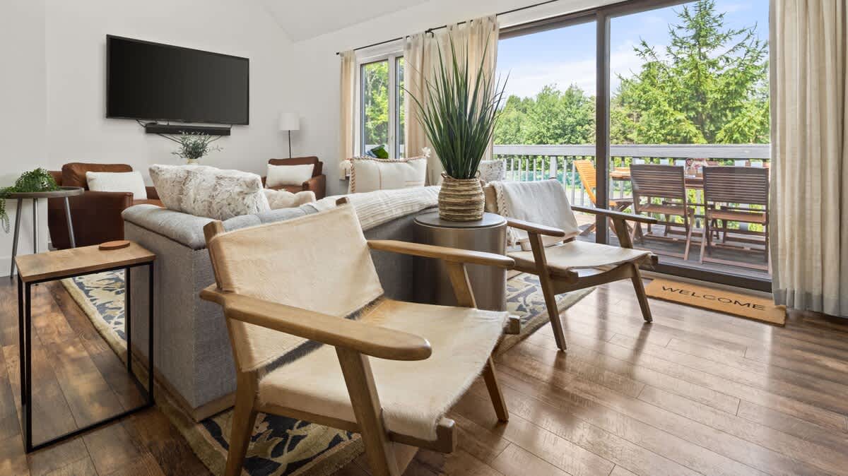 Bright living room with a gray sofa and two light-colored sling chairs, facing large sliding glass doors that open to a deck with outdoor seating and lush green trees. A wall-mounted TV is visible in the background, and a patterned rug anchors the seating area.