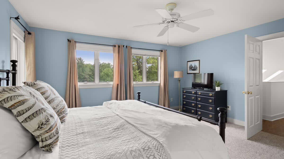 A bright bedroom with light blue walls, a black metal bed frame with white bedding, and two windows draped with light brown curtains overlooking green trees. A ceiling fan hangs above, and a dark dresser with a TV is visible.
