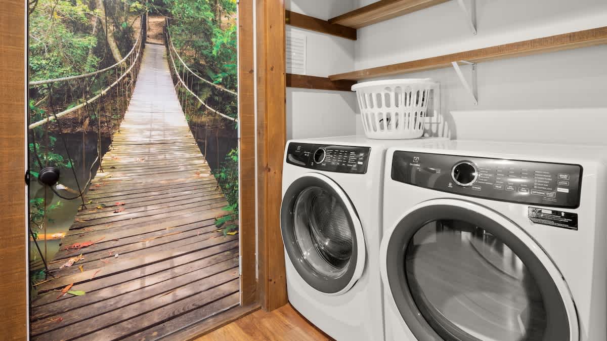 A dedicated laundry area with a white front-loading washer and dryer set, wooden shelving above, and a white laundry basket. A door with a vibrant mural of a suspension bridge in a jungle adds a unique touch.