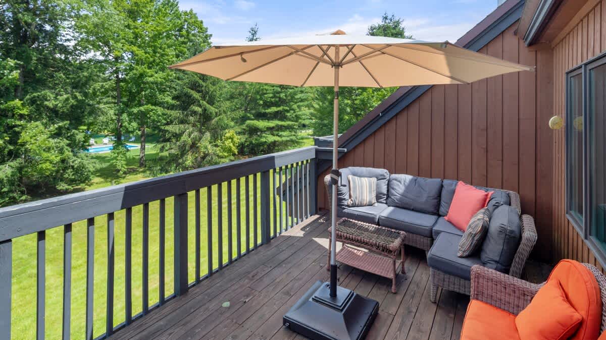 A spacious wooden deck with dark gray railings, furnished with a gray outdoor sectional sofa, a wicker coffee table, and an open beige patio umbrella. The deck overlooks a vibrant green lawn and tall trees.