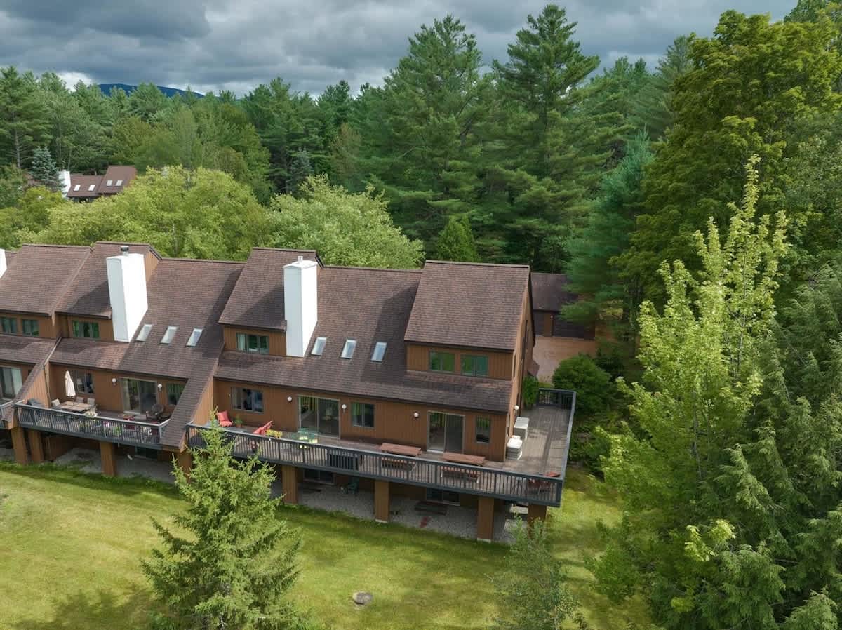 An aerial view of a multi-unit condominium building with brown siding and dark brown roofs, nestled among dense green trees. Multiple decks are visible on the back of the units, overlooking a grassy area.