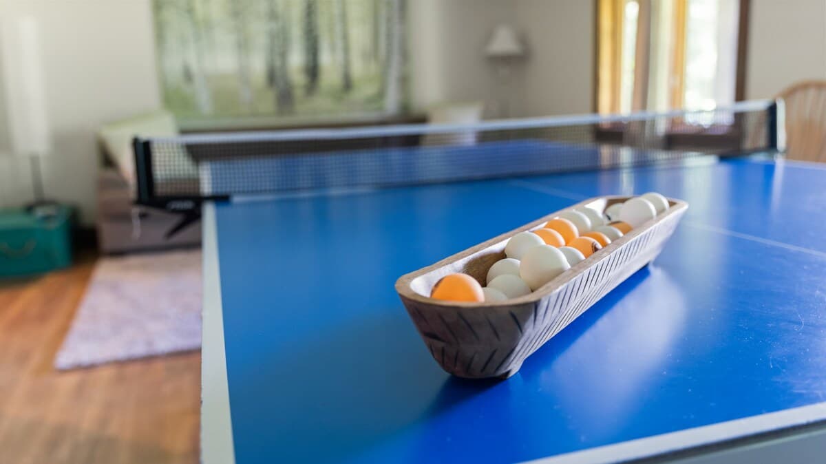 A close-up view of a blue ping-pong table with a net, featuring a wooden bowl filled with white and orange ping-pong balls. The background shows a blurred living area with a sofa and artwork.