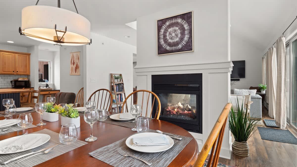 Elegant dining area with a round wooden table set for guests, illuminated by a modern drum pendant light. A cozy fireplace with a white mantel separates the dining space from the living room, and a kitchen with light wood cabinets is visible in the background.