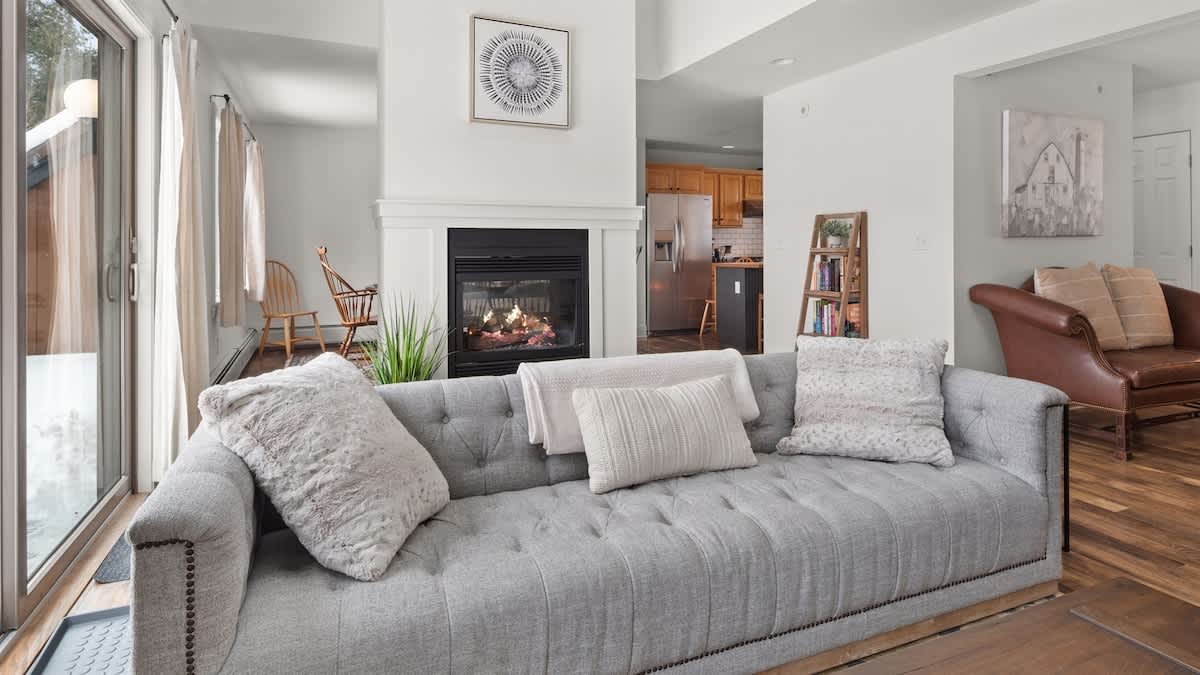 A bright living room with light hardwood floors, a comfortable grey tufted sofa with throw pillows, and a modern white fireplace feature separating it from the dining area.