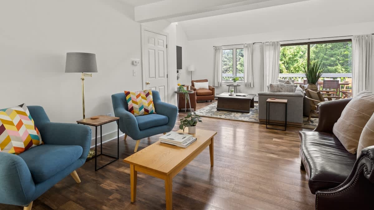 Bright living room with light walls, dark hardwood floors, and a mix of blue armchairs and a dark leather sofa, centered around a light wood coffee table. Large sliding glass doors lead to an outdoor deck with lush greenery.