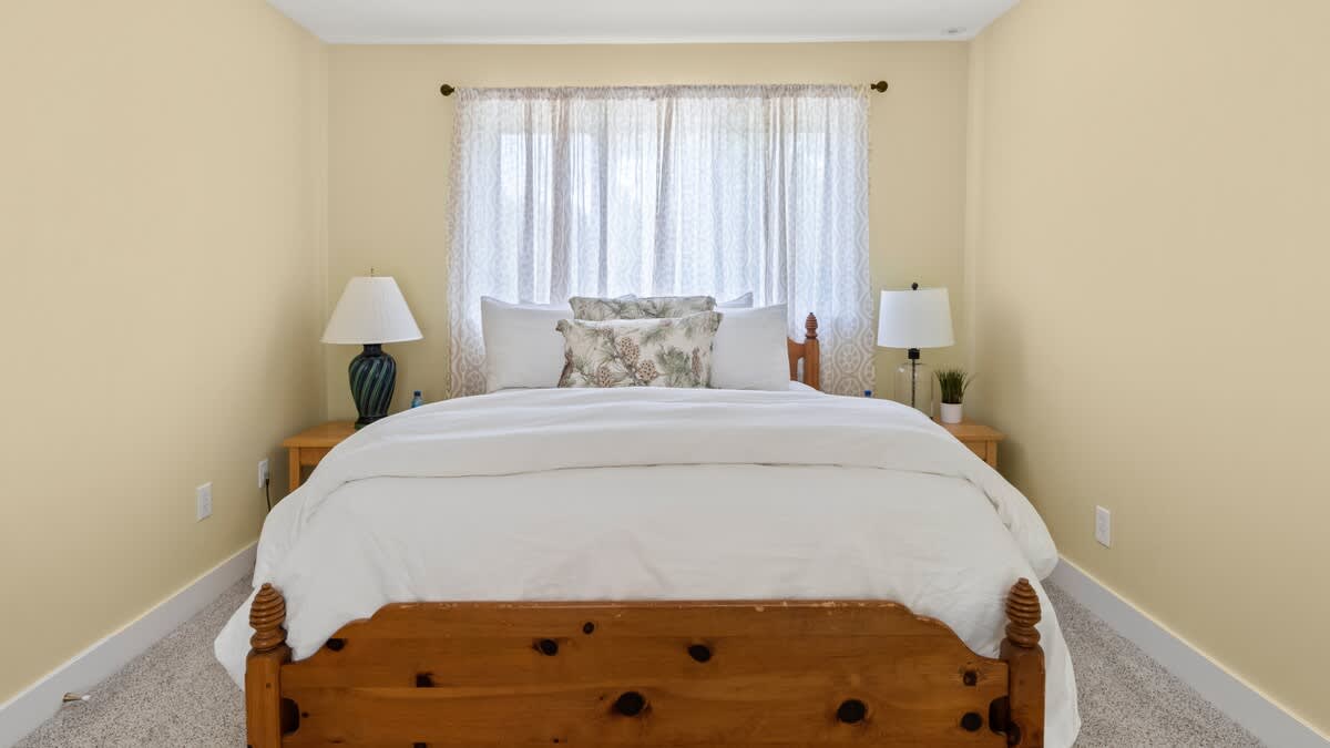 A cozy bedroom with light yellow walls and a rustic wooden bed frame dressed in white bedding and decorative pillows. A large window with sheer white curtains allows natural light, flanked by two wooden nightstands with lamps.