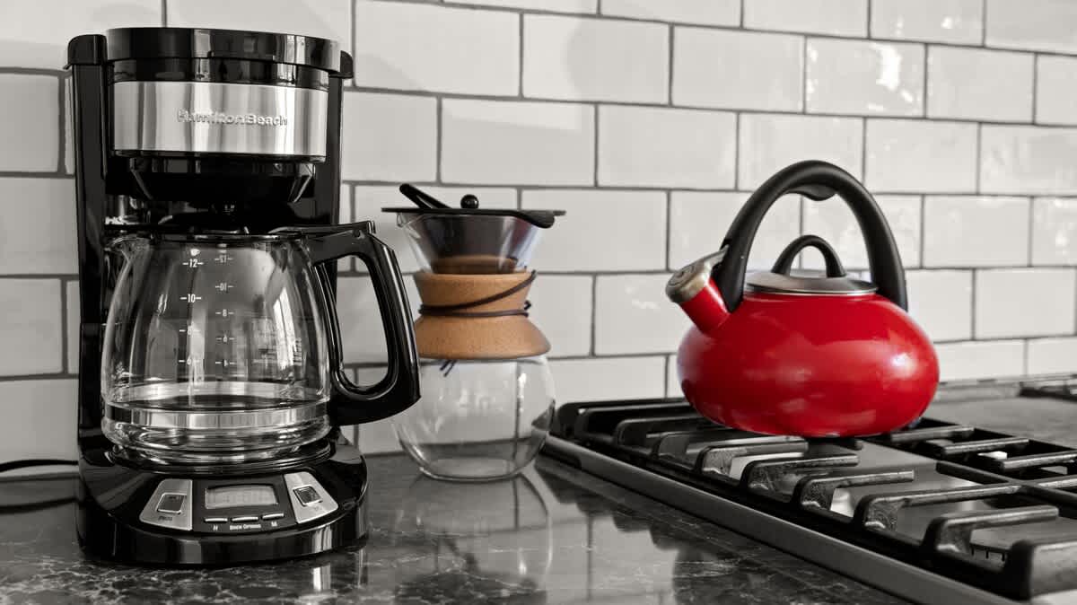 Kitchen counter detail showing a black drip coffee maker, a pour-over coffee setup, and a vibrant red whistling kettle on a gas stovetop, all against a white subway tile backsplash.