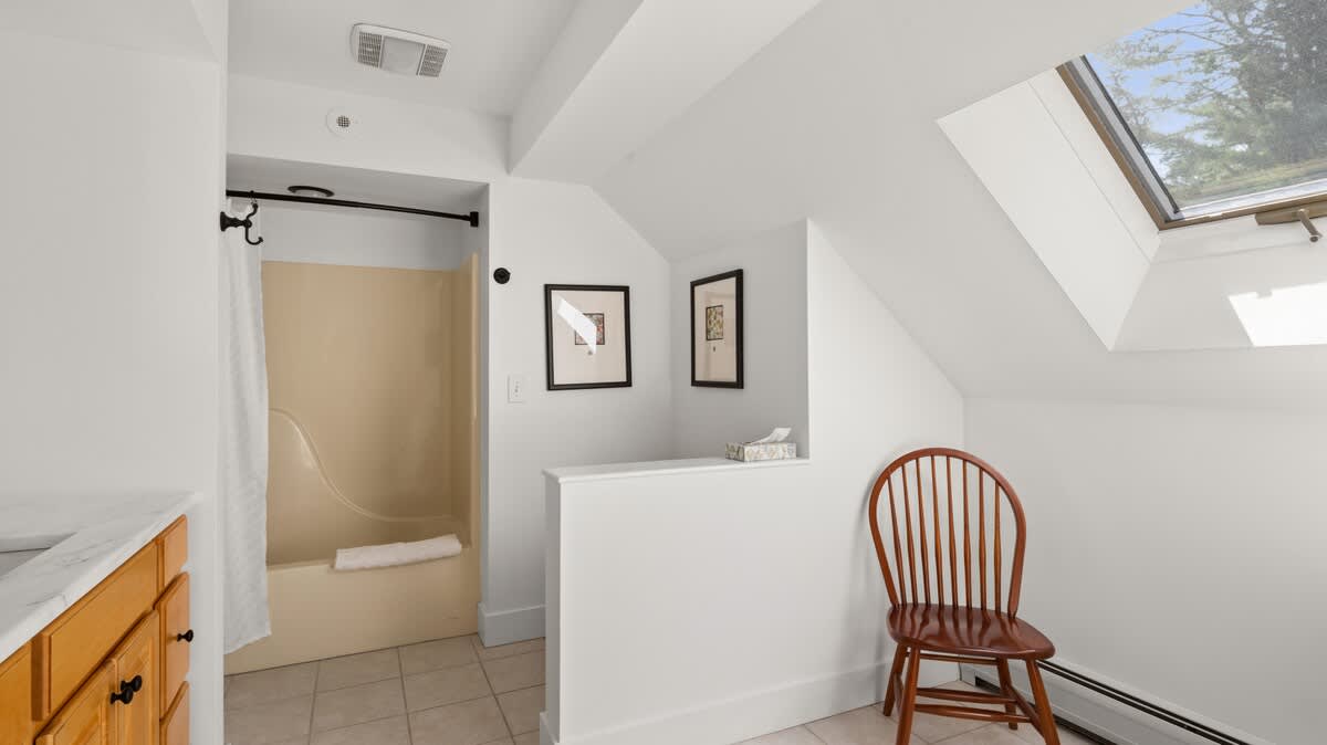 A bright bathroom with white walls and a sloped ceiling featuring a skylight. It includes a beige tub/shower combo, a wooden vanity with a sink, and a wooden chair.