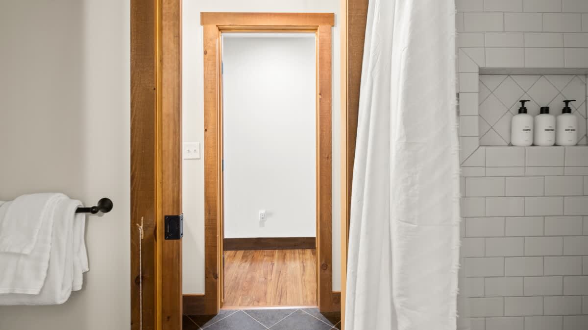 A clean and modern shower area featuring white subway tiles and a built-in niche for toiletries, separated by a white shower curtain. The dark tiled floor matches the adjacent bathroom area, and a wooden door frame is visible.