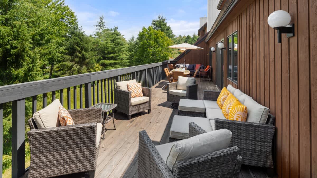 An expansive wooden deck furnished with multiple outdoor seating arrangements, including wicker chairs with light cushions and yellow throw pillows, and a dining area with an umbrella. The deck is surrounded by lush green trees.