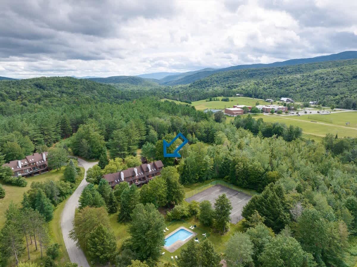 A wide aerial shot showcasing a condominium complex surrounded by lush green forests and distant mountains under a cloudy sky. A tennis court and a swimming pool are visible nearby, with an arrow pointing to the specific unit.