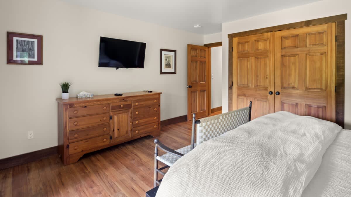 A warm and inviting bedroom featuring light beige walls, rich wooden floors, and a large wooden dresser with a wall-mounted TV. A bed with white linens is partially visible, and wooden bi-fold doors lead to a closet.