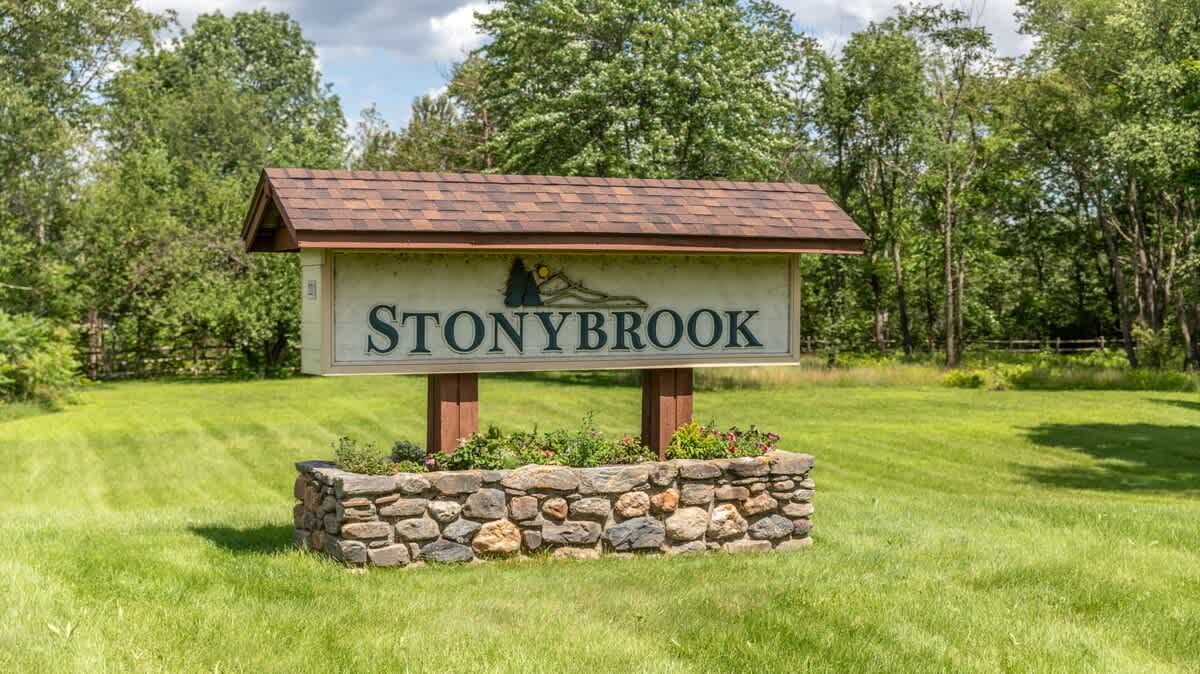 A rustic wooden sign for "STONYBROOK" with a small roof, set on a stone base filled with plants, surrounded by a well-maintained green lawn and trees in the background.