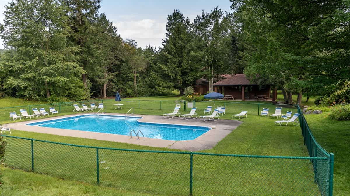 A rectangular outdoor swimming pool with blue water, surrounded by a green chain-link fence and white lounge chairs. A small building with a covered picnic area is visible in the background, all nestled among tall trees.
