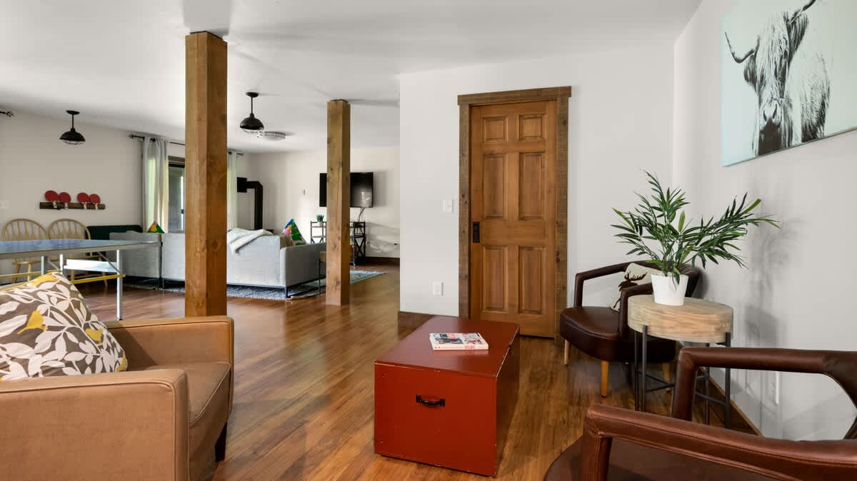 An open-concept living area with warm hardwood floors, featuring a brown armchair, a red trunk coffee table, and a wooden door. Exposed wooden pillars add a rustic touch, and a large framed artwork of a highland cow hangs on the wall.