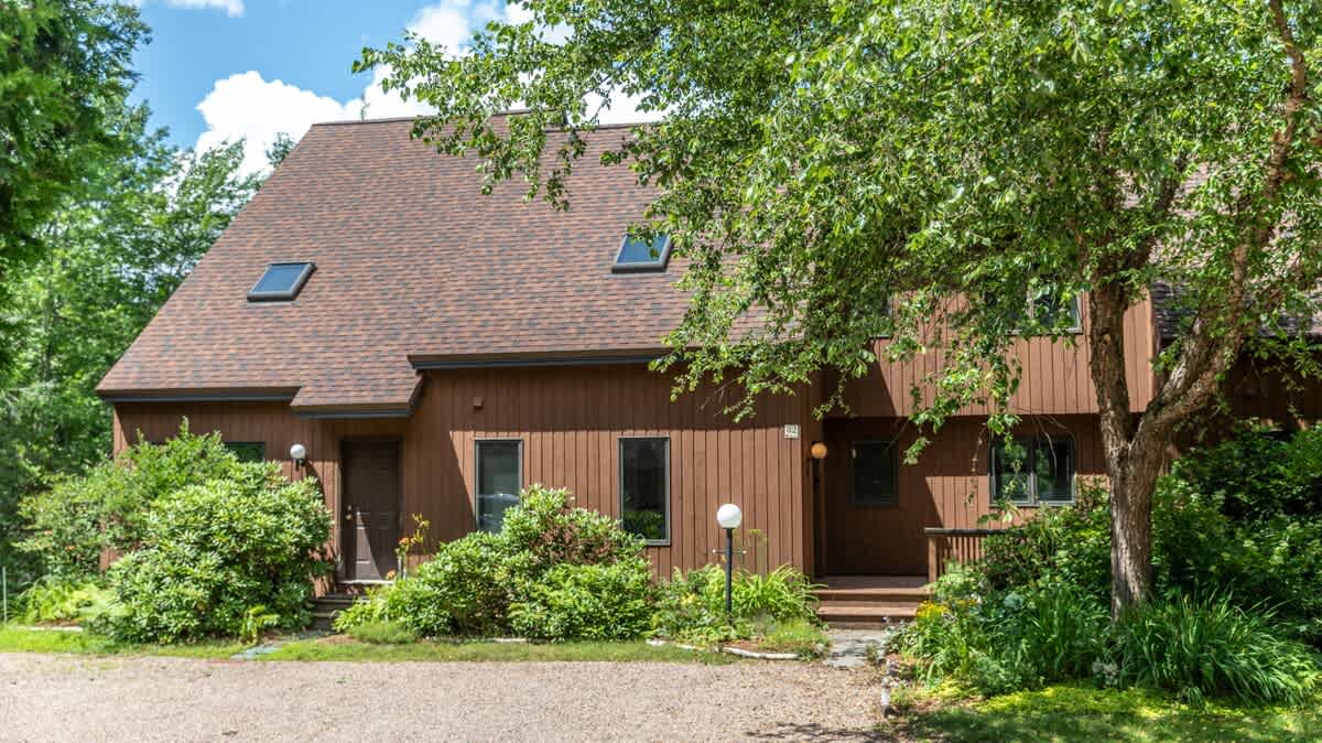 A brown, multi-level condominium building with a dark shingle roof and skylights, surrounded by lush green trees and bushes under a bright blue sky. A gravel driveway leads to the front entrances.