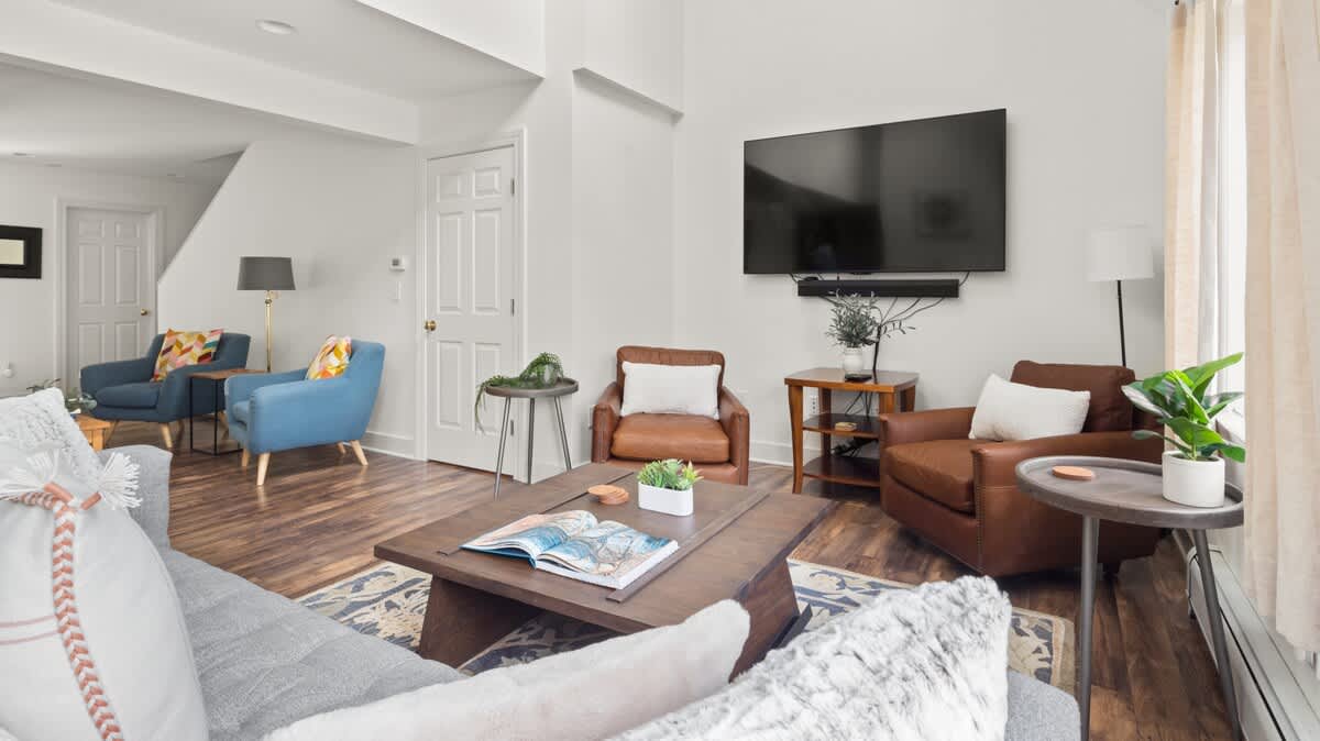 Modern living room featuring a wall-mounted TV above a soundbar, two brown leather armchairs, and a gray sofa, all arranged on a patterned rug over dark hardwood floors. A white door and staircase are visible in the background.