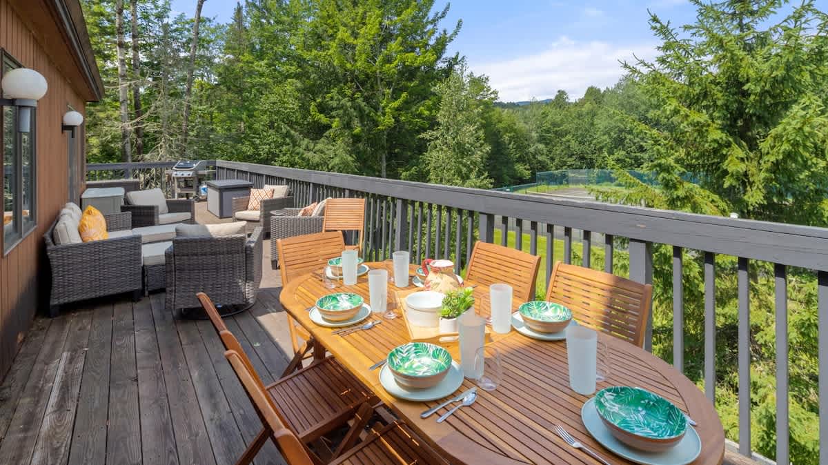 A large outdoor wooden deck furnished with a long dining table set for six, and a separate lounge area with wicker seating, all overlooking a lush green landscape and tennis courts.