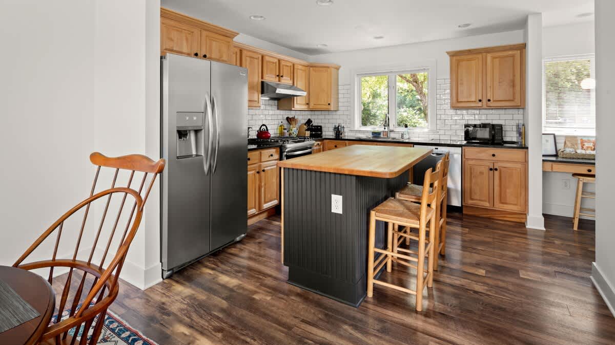 A spacious kitchen with light wood cabinetry, stainless steel appliances, a white subway tile backsplash, and a dark island with a butcher block countertop and bar seating.