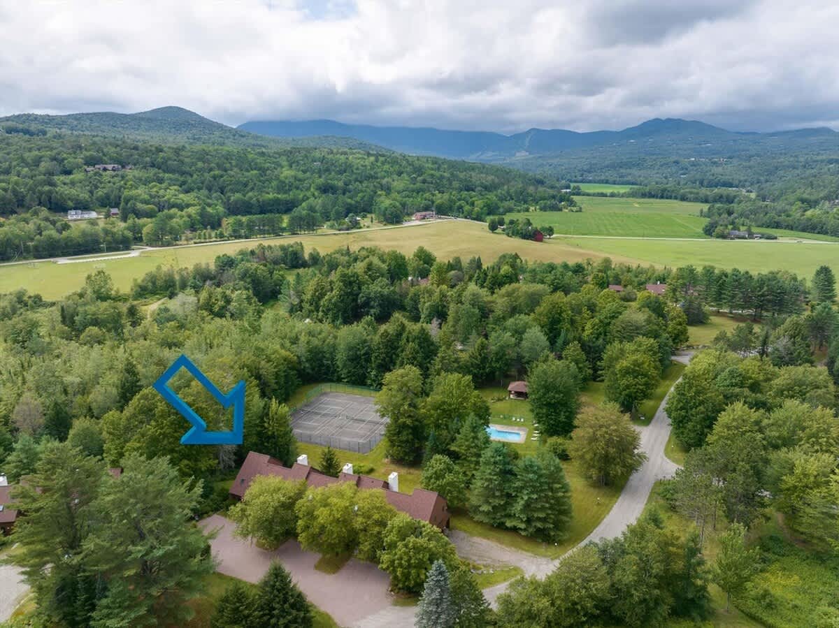 An aerial perspective of the Stonybrook condominium complex, highlighting its proximity to a tennis court and a swimming pool, all set within a dense forest landscape with rolling mountains in the distance. An arrow indicates the specific unit.
