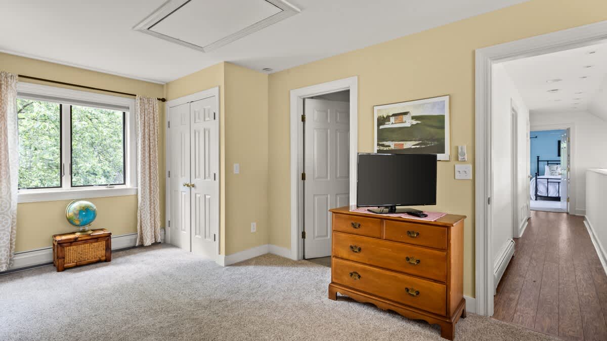 A bright room with light yellow walls and a large window with patterned curtains. A wooden dresser with a television sits against one wall, and a globe on a small wooden trunk is by the window.