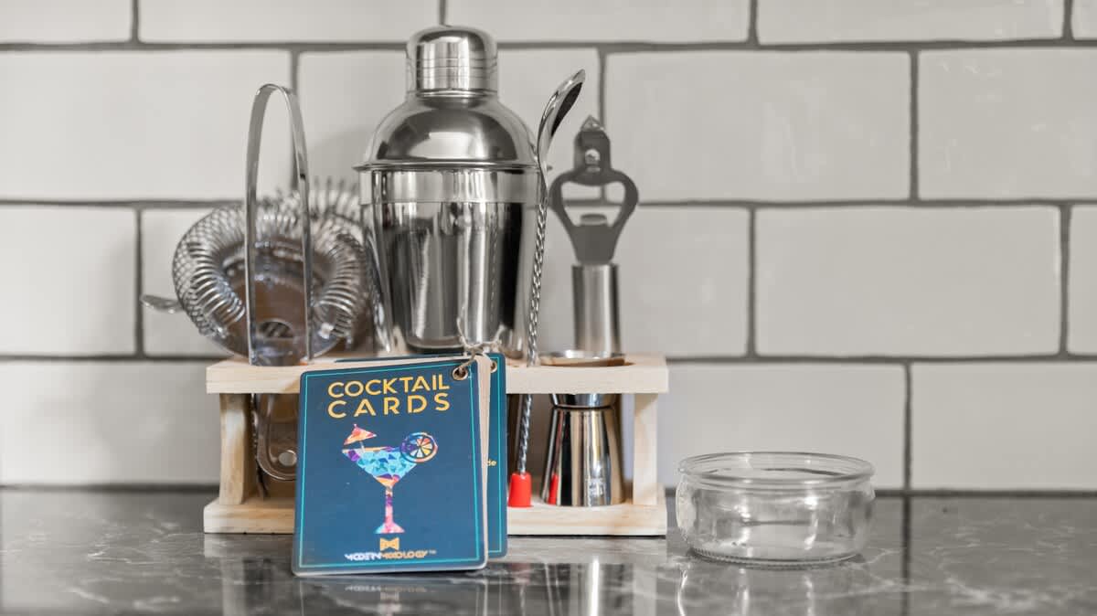 Close-up of a bar cart setup on a black countertop against a white subway tile backsplash, featuring a stainless steel cocktail shaker set and a stack of "Cocktail Cards."