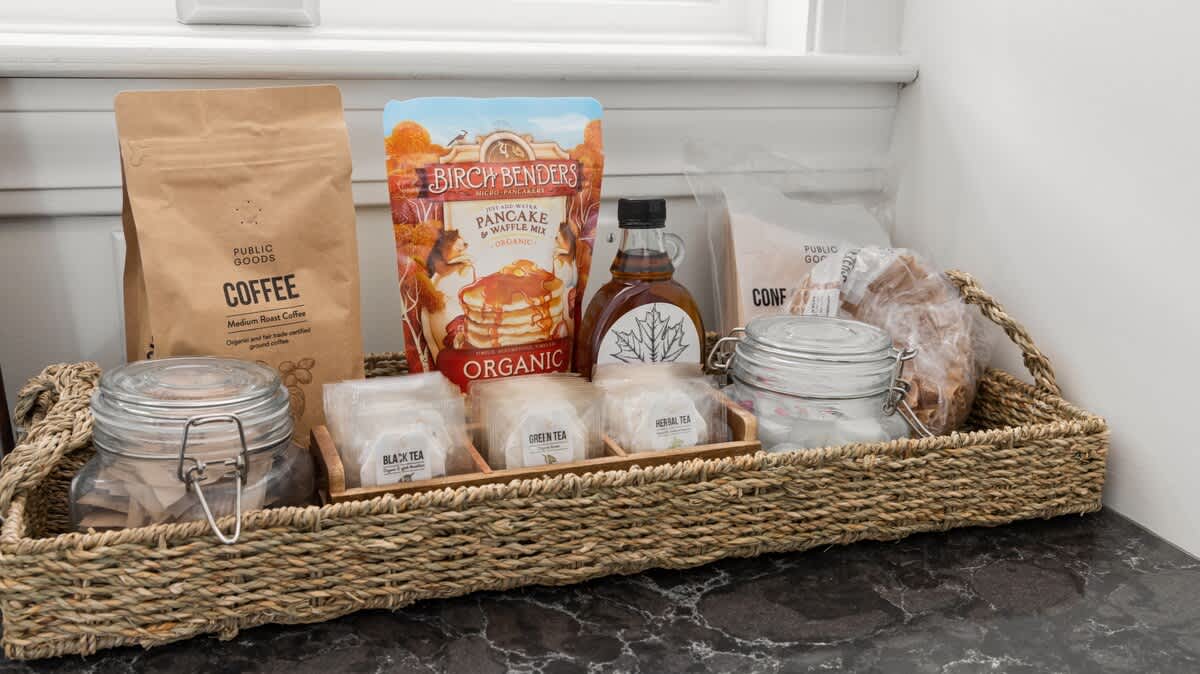 A close-up of a woven tray filled with breakfast essentials, including coffee, pancake mix, maple syrup, various teas, and what appears to be granola or pastries, set on a dark countertop.