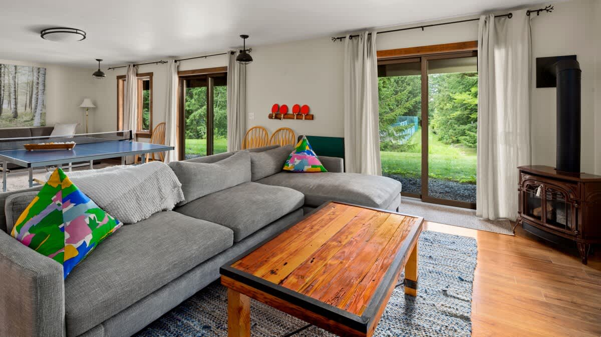A spacious living room featuring a large gray sectional sofa with colorful throw pillows, a rustic wooden coffee table on a blue patterned rug, and a ping pong table in the background. Large sliding glass doors offer views of lush greenery.