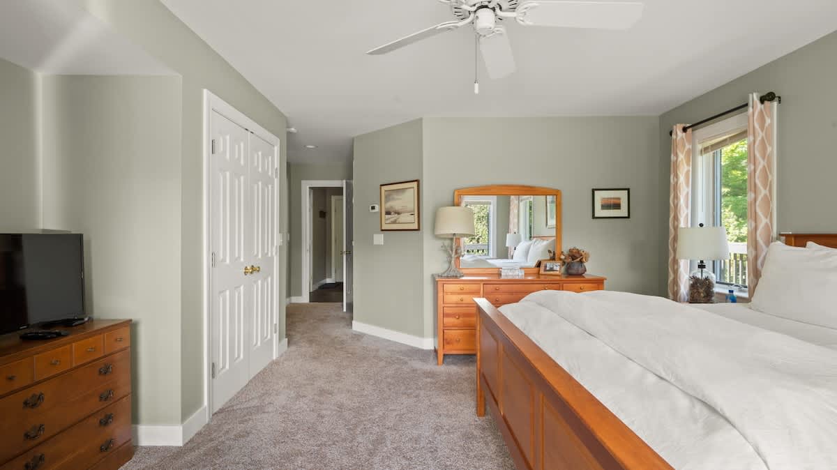 A spacious bedroom featuring sage green walls, a wooden bed frame with white bedding, and a large wooden dresser with a mirror. Two windows with patterned curtains offer views of greenery, and a ceiling fan is mounted overhead.