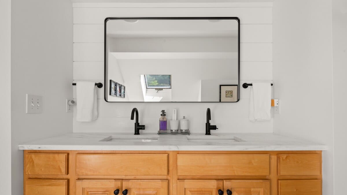 A close-up of the double vanity in a modern bathroom, showcasing a light wood cabinet, white marble countertop, and two black-finished faucets. A large rectangular mirror with a black frame is mounted above, reflecting the room.