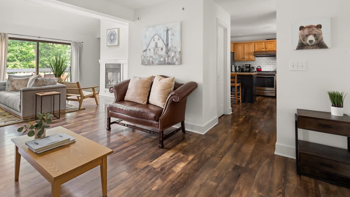 Spacious living area with dark hardwood floors, a brown leather settee, and a light wood coffee table, leading into a kitchen with light wood cabinets and a dining area. A large painting of a barn hangs on the wall, and a bear portrait is visible further down the hall.