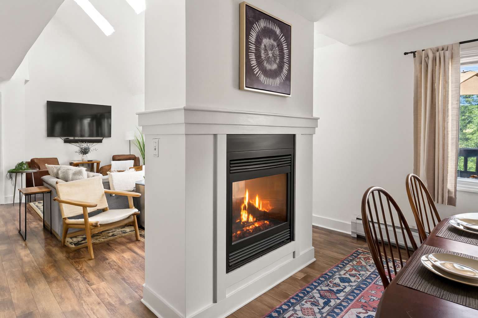 A modern living area with light hardwood floors and a white fireplace feature, separating the seating area with a large TV from a dining space with a wooden table and chairs.