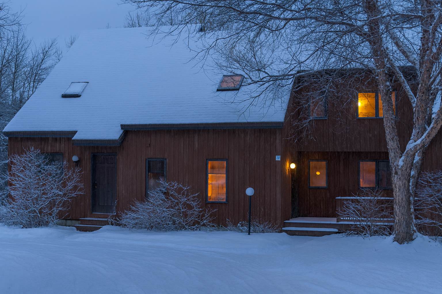 A two-story, brown-sided house with a snow-covered roof and ground, illuminated by warm interior lights from several windows, surrounded by bare trees and snow-dusted bushes under a twilight sky.