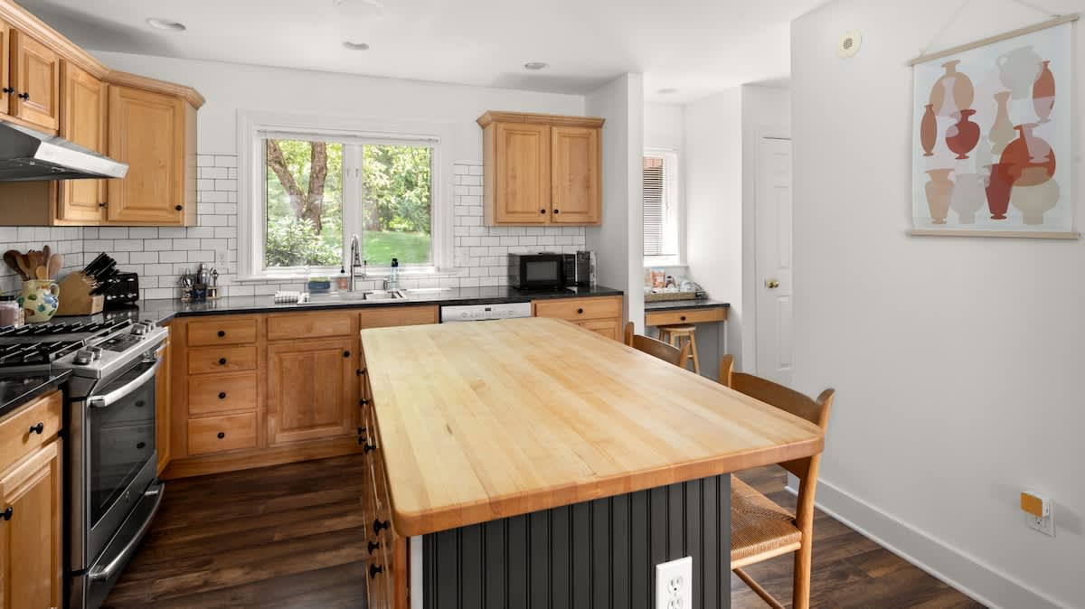 Well-lit kitchen featuring light wood shaker cabinets, black countertops, and a white subway tile backsplash. A large wooden island with a butcher block top and black base provides additional workspace, with a window overlooking greenery.