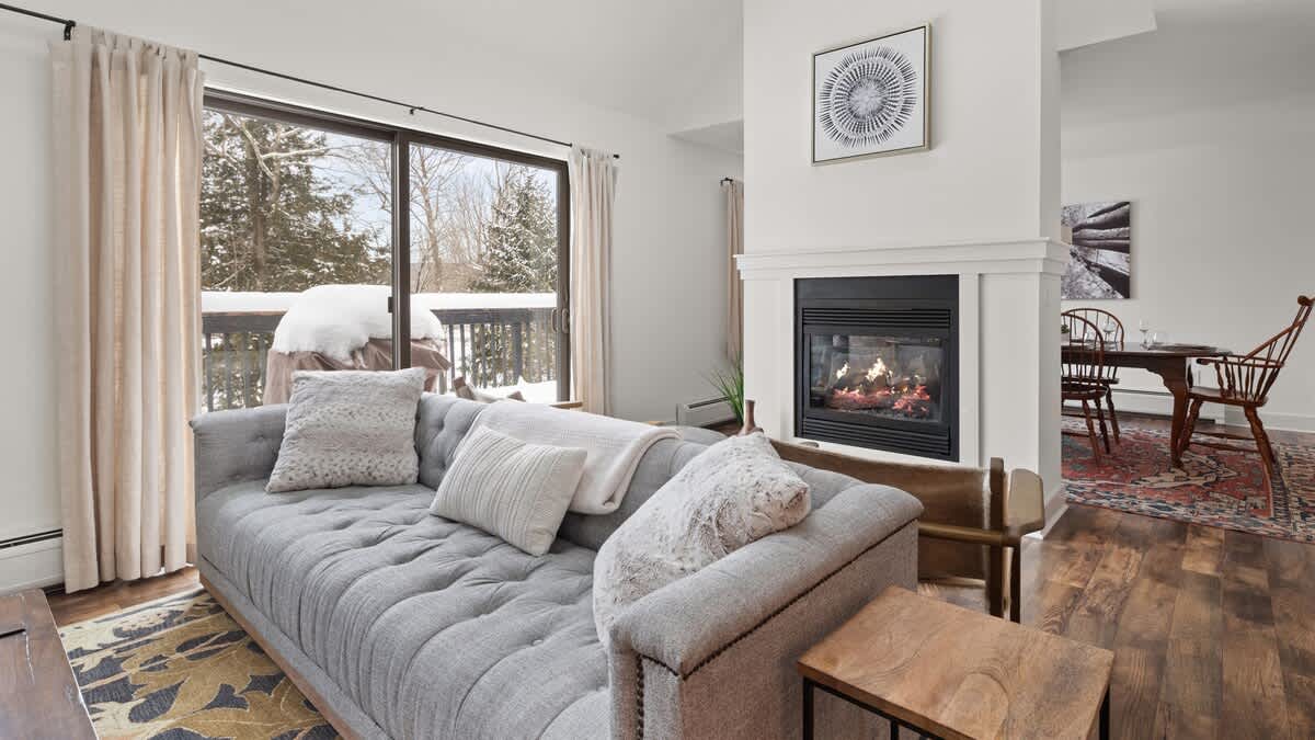 Cozy living room with a gray tufted sofa facing a modern fireplace with a white mantel, and large sliding glass doors opening to a snowy outdoor deck. A patterned rug covers dark hardwood floors, and a dining area is visible to the right.