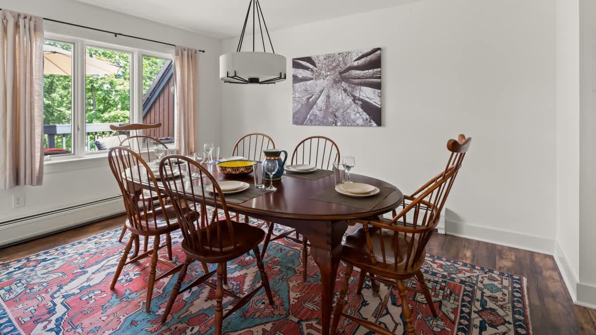 A cozy dining room with a dark oval wooden table surrounded by six wooden spindle-back chairs, set on a vibrant patterned rug. A modern chandelier hangs above, and a large window offers a view of the outdoors.