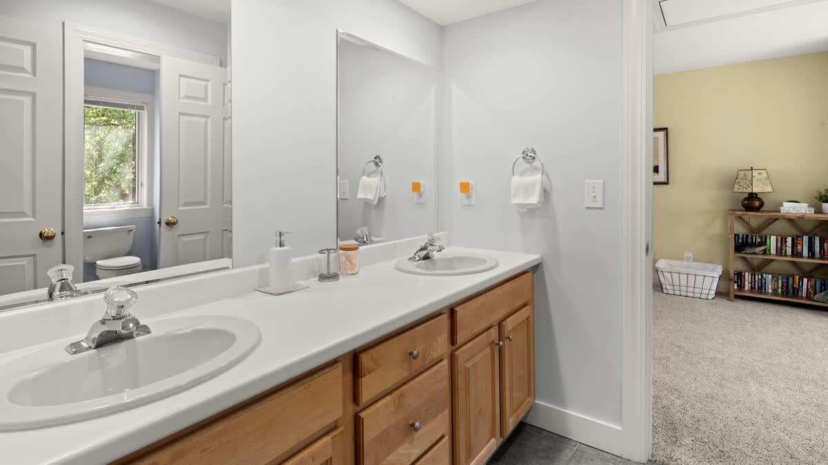 A modern bathroom featuring a light wood double vanity with two sinks and a large mirror. White walls and a grey tiled floor complete the space, with a view into an adjacent room.