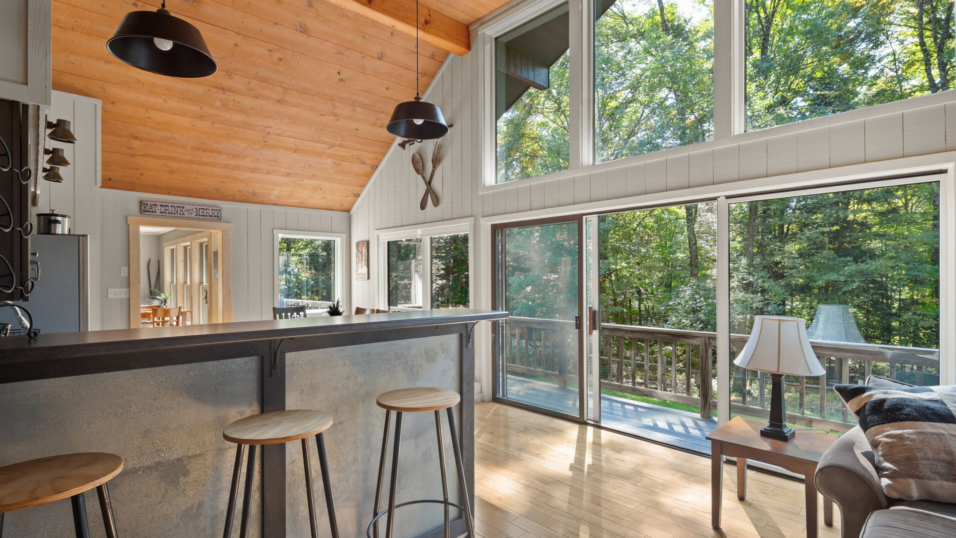 Bright open-concept living area with light wood floors and a vaulted wood-paneled ceiling, featuring a bar with stools and large sliding glass doors leading to an outdoor deck. Expansive windows offer views of the surrounding lush forest.