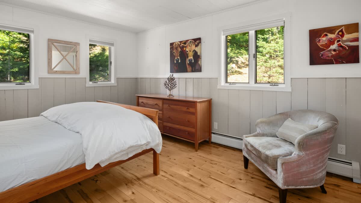 A bright bedroom with light wood floors, white shiplap walls, and a wooden bed frame with white bedding. A wooden dresser, a velvet armchair, and two windows offering views of lush greenery complete the space.