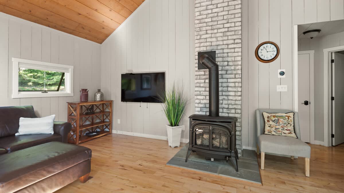 Living room area with light wood floors and light gray shiplap walls, featuring a wall-mounted flat-screen TV and a black wood-burning stove on a stone hearth. A brown leather sofa and a wooden console table are also present.