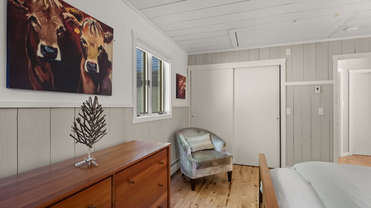 A bright bedroom with light wood floors and white shiplap walls, featuring a wooden dresser with cow artwork above it. A velvet armchair sits beside a window, and a sliding closet door is visible.