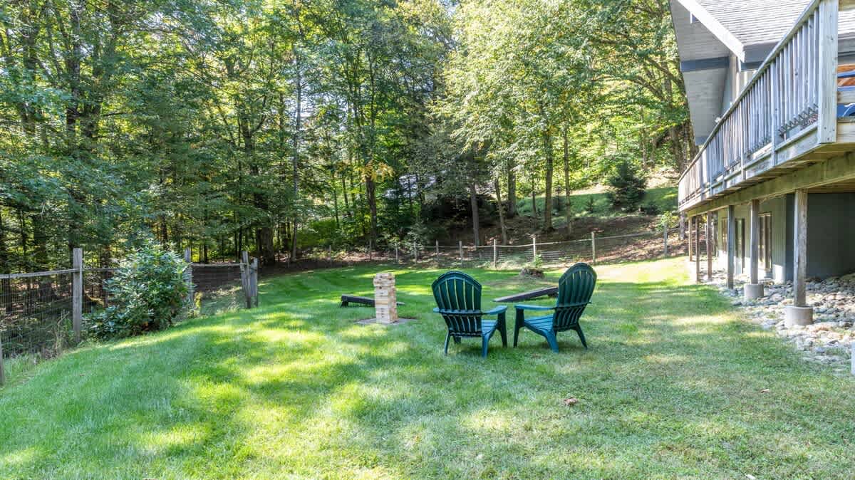 Expansive green backyard with two dark green Adirondack chairs and a stone fire pit area, bordered by a fence and mature trees, offering a peaceful outdoor retreat.