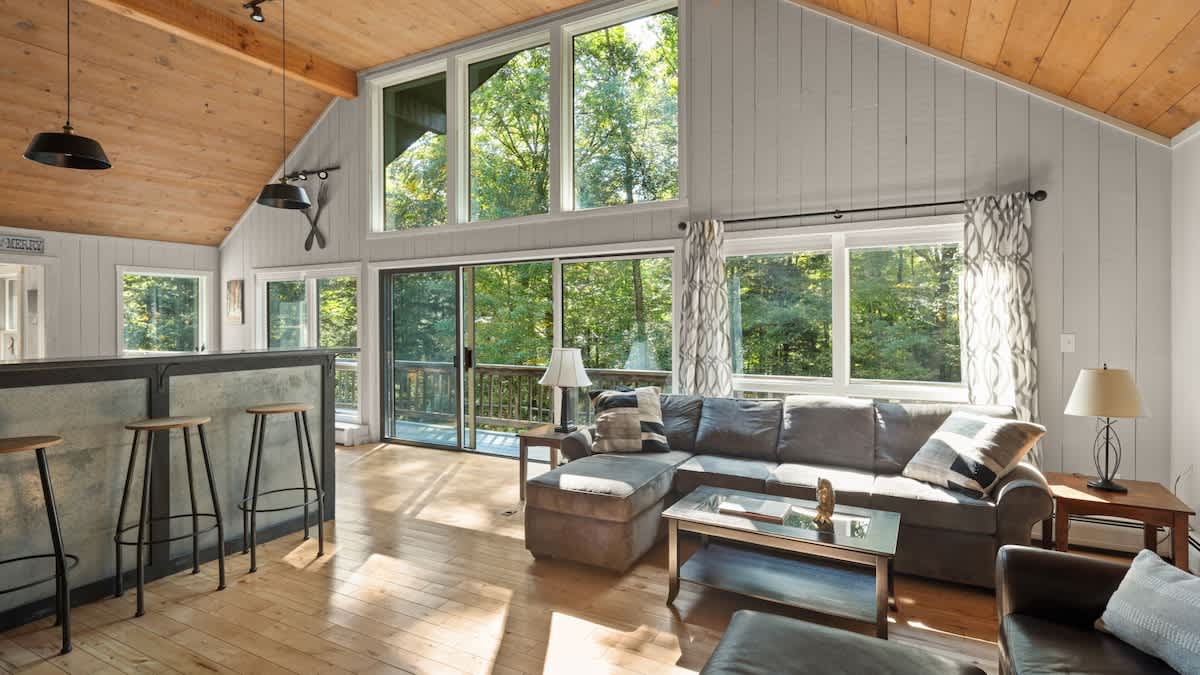 Cozy living room with light wood floors, a vaulted wood-paneled ceiling, and large windows offering expansive views of the surrounding trees. A gray sectional sofa and a coffee table are arranged, with a bar area visible to the left.