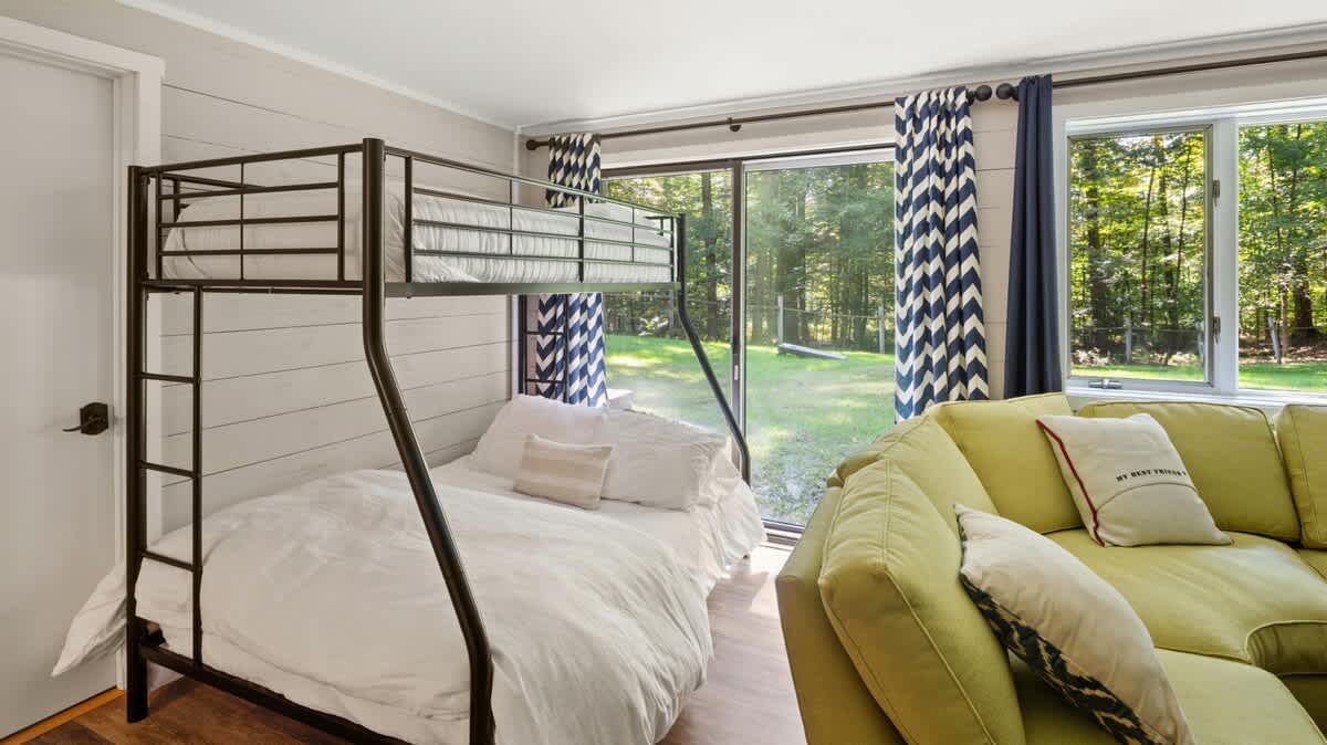 A metal bunk bed with white bedding is positioned next to large windows with chevron-patterned curtains, offering a view of the lush green backyard and surrounding trees.