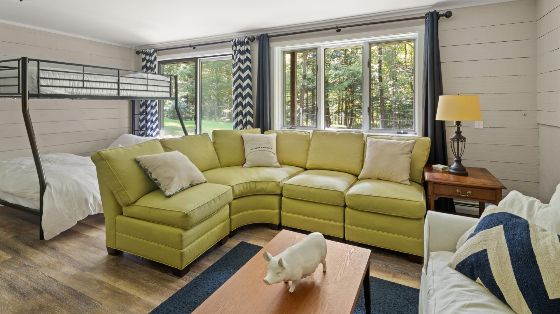 Bright living room with a large lime green sectional sofa, a white ceramic pig figurine on a wooden coffee table, and a metal bunk bed visible near large windows overlooking a wooded area.