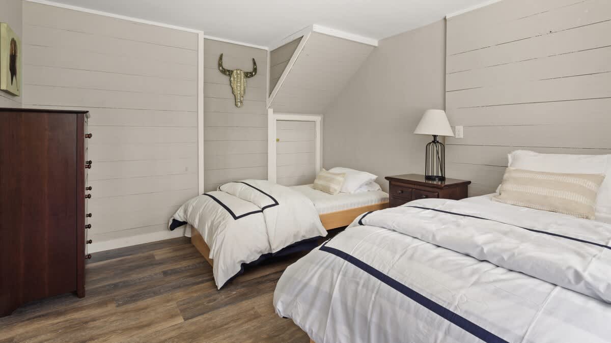 A rustic bedroom with two twin beds, dark wood-look flooring, and light gray shiplap walls. A tall wooden dresser and a bull skull wall decor are visible, with an angled ceiling adding character.