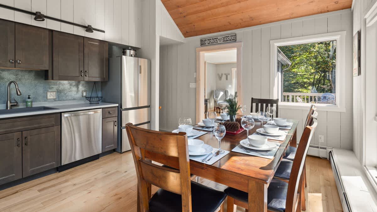 Bright dining area with a wooden table set for six, surrounded by dark wood chairs, situated next to a large window overlooking the outdoors. The kitchen cabinets and stainless steel appliances are visible on the left, blending into the open-concept space.