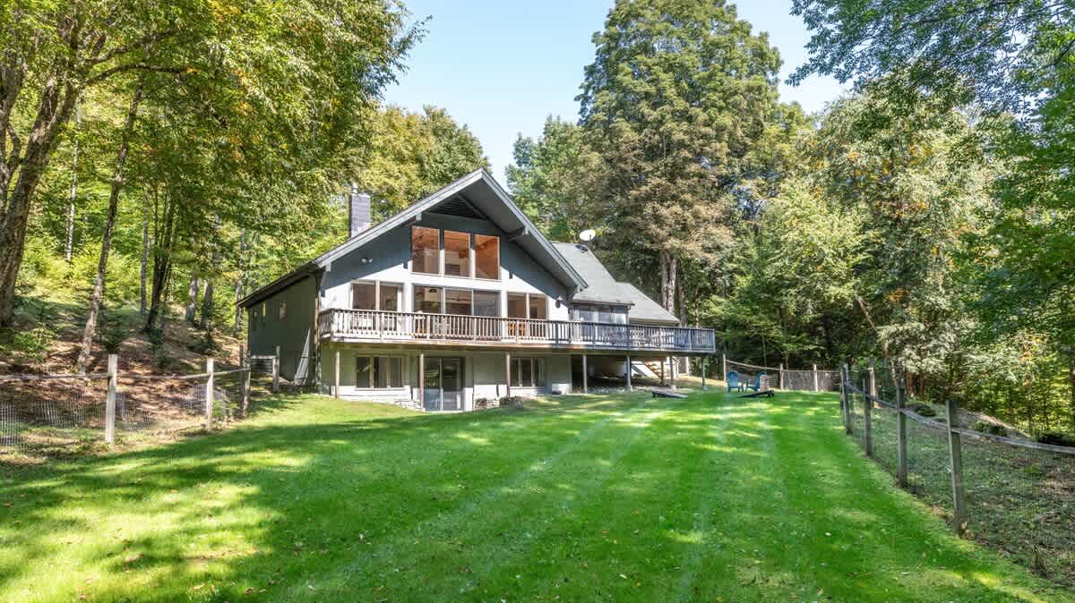 Stunning back exterior of the house, featuring a large deck, expansive windows, and a walk-out lower level, all set against a backdrop of a vast green lawn and dense forest.