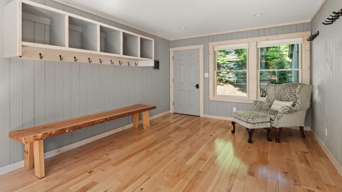 Functional mudroom with light wood floors and light gray vertical shiplap walls, featuring a long wooden bench and a built-in cubby shelf with hooks for coats and gear. A window and a light gray door are also visible.