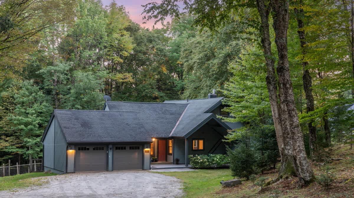 Charming dark gray chalet-style house with a multi-gabled roof and attached two-car garage, illuminated at dusk and surrounded by tall, mature trees in a serene wooded environment.